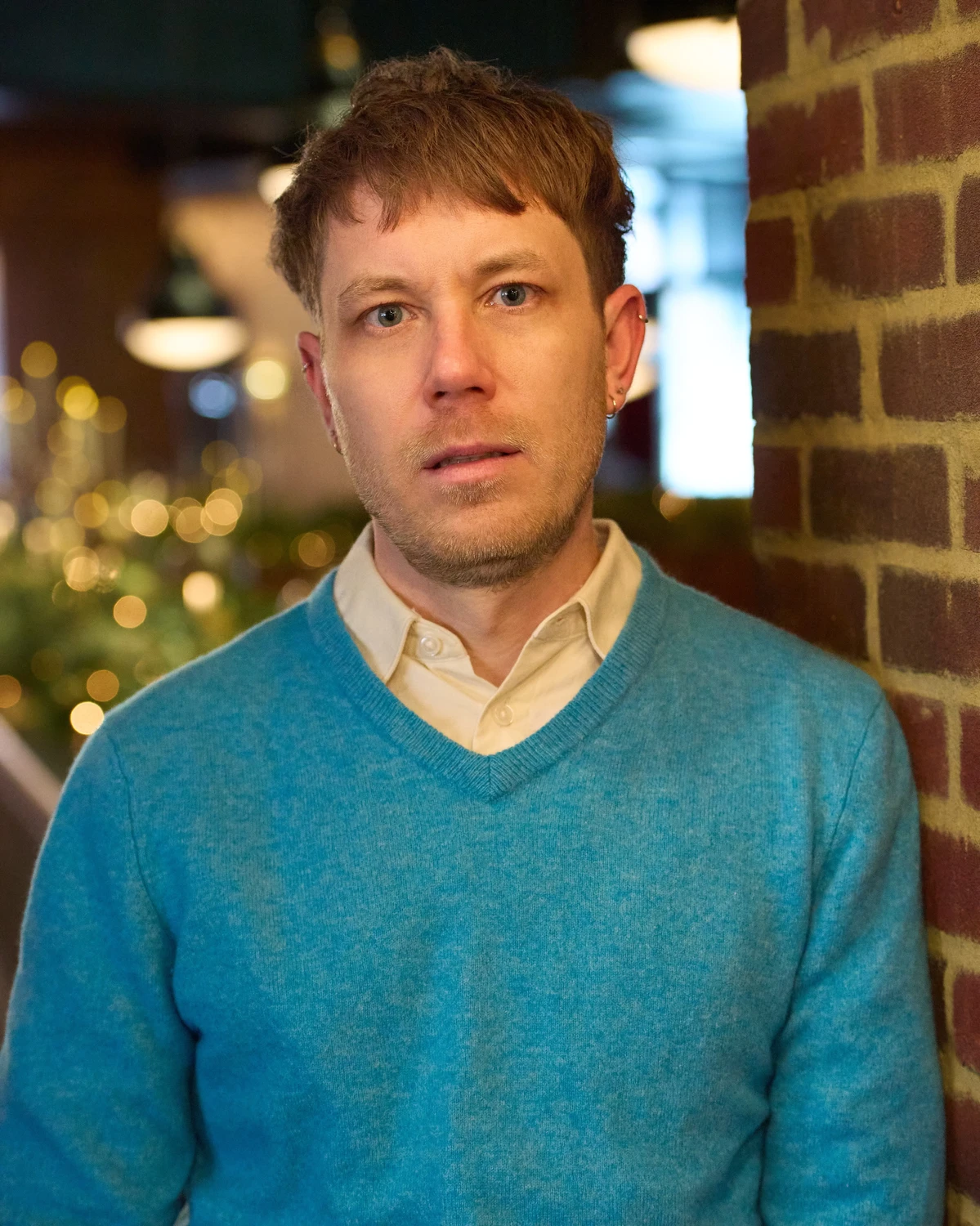 Stephen Kochenash, A holistic, relational therapy practice on the border of Nomad and Chelsea. Wearing a blue shirt and standing in front of a brick wall to the right and blurry plants and string lights to the left.